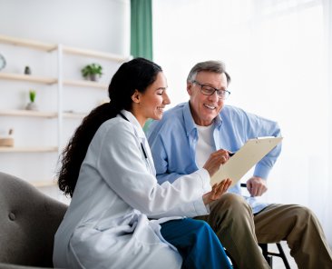 Young doctor asking senior impaired male patient in wheelchair to sign insurance policy at home. Handicapped elderly man putting his signature under surgery consent form, reading medical document